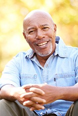 Man in denim shirt outside smiling with hands clasped